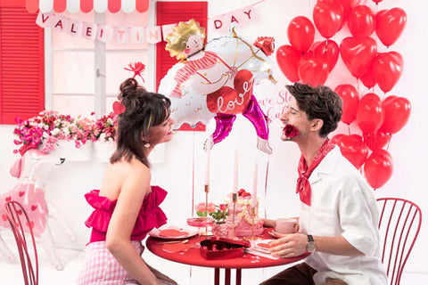 Two people at a Valentine's Day table with Cupid foil balloon and red heart balloons in romantic setting