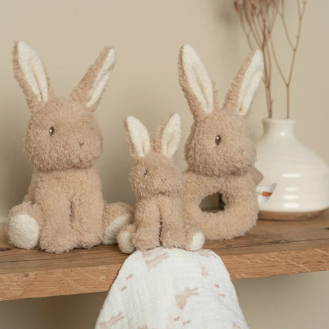 „Little Dutch Baby Bunny“ pliušinis kiškis, baltas migdukas su kiškučio galvute ir žiedinis barškutis ant medinės lentynos.